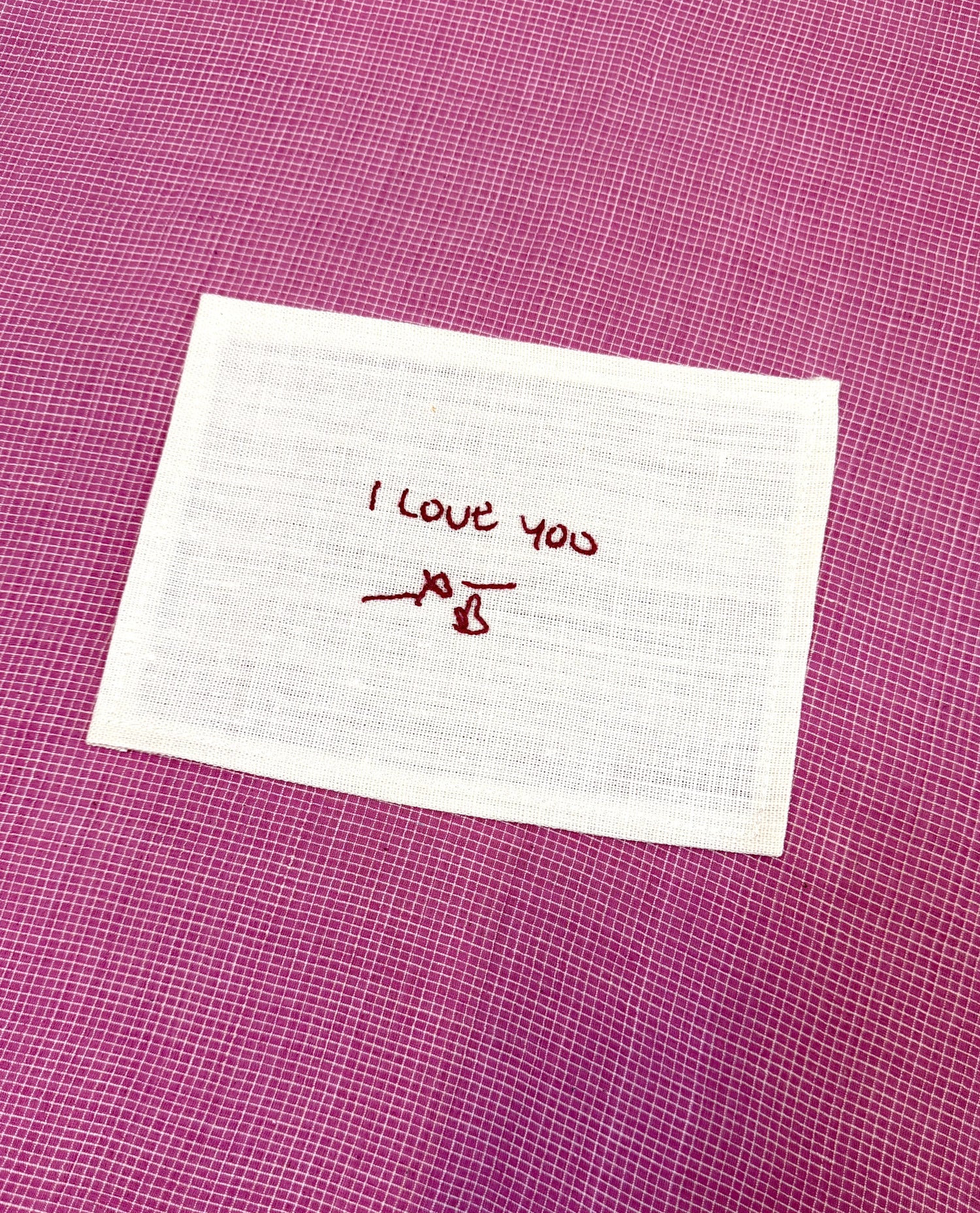 A square hand embroidered fabric love note with the words 'I love you' stitched in red thread, placed on a textured magenta background.